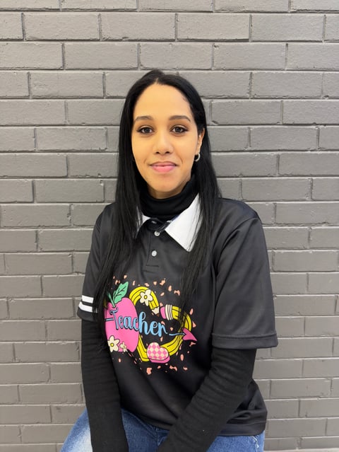 Woman with long black hair wearing a black graphic tee and white collared shirt, posing in front of a gray brick wall