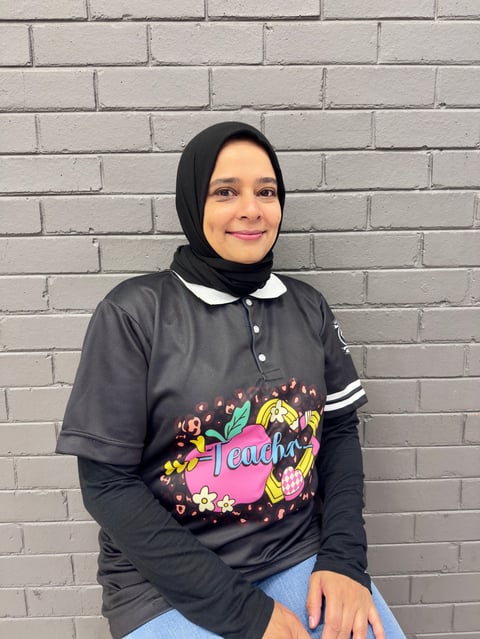 Young woman wearing a black hijab and black graphic t-shirt over a long-sleeve shirt, smiling against a white brick wall