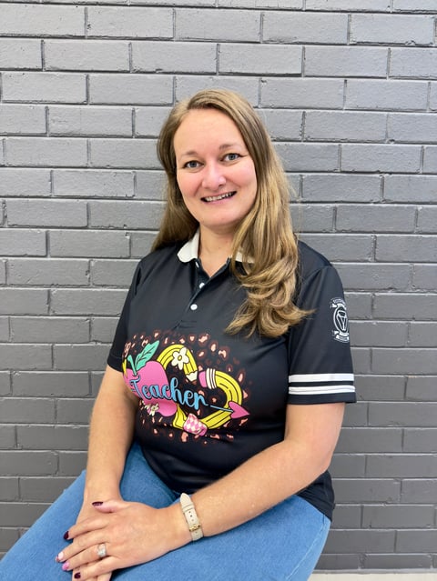Smiling woman with long blonde hair wearing a black polo shirt with colorful graphics, seated against a gray brick wall