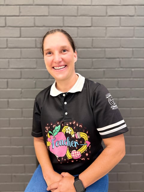 Woman smiling at camera wearing black polo shirt with teacher graphic and striped sleeves against gray brick wall