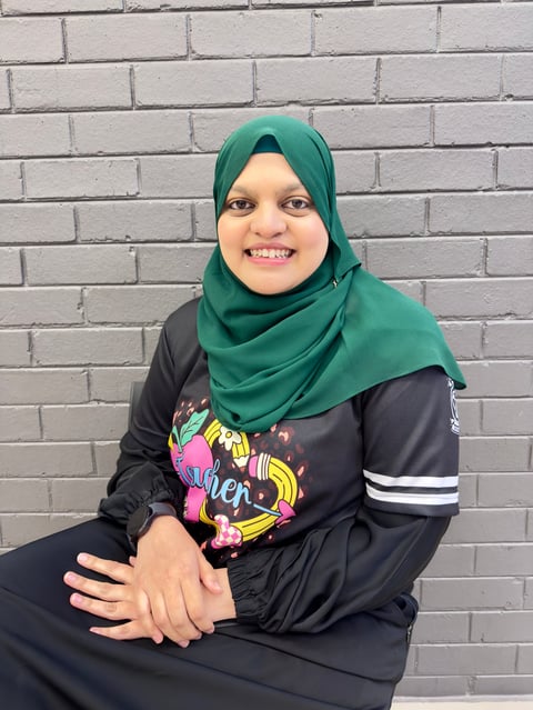 Young woman wearing a green hijab and black graphic hoodie sitting against a brick wall, smiling at the camera