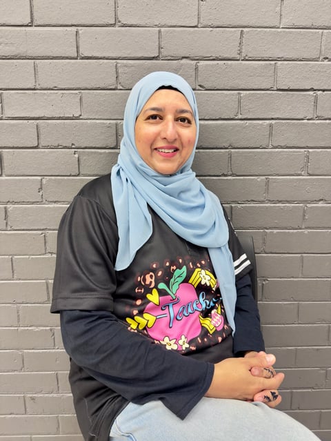 Woman wearing light blue hijab and dark graphic t-shirt smiling at camera against light brick wall