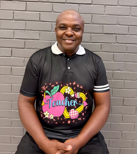 Smiling man wearing a black polo shirt with colorful Teacher graphic print, seated against a gray brick wall