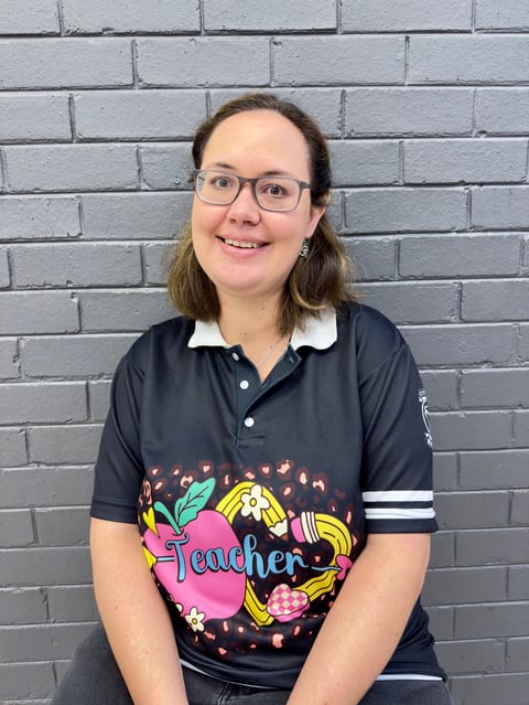Woman with glasses smiling at camera wearing a navy teacher-themed polo shirt against a brick wall