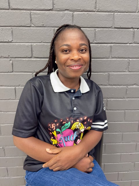 Smiling woman with braided hair wearing a dark polo shirt with colorful graphic design and jeans, posing against a gray brick wall