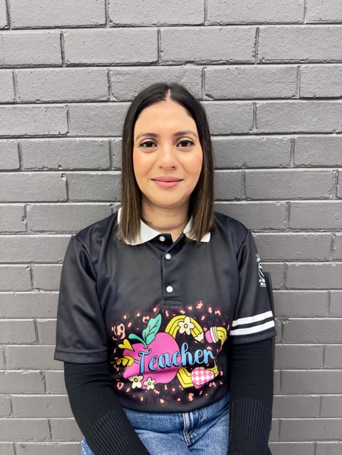 Woman with long dark hair wearing a black and colorful teacher graphic shirt against a white brick wall