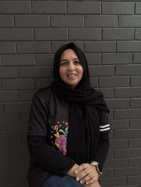 Young woman wearing a black hijab and graphic hoodie smiling at camera against a gray brick wall