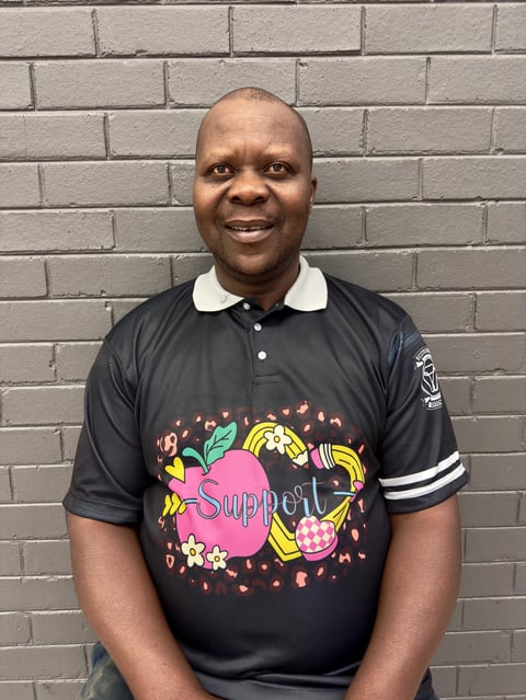 Smiling man wearing black polo shirt with colorful fruit graphic and white collar stripes, standing in front of gray brick wall