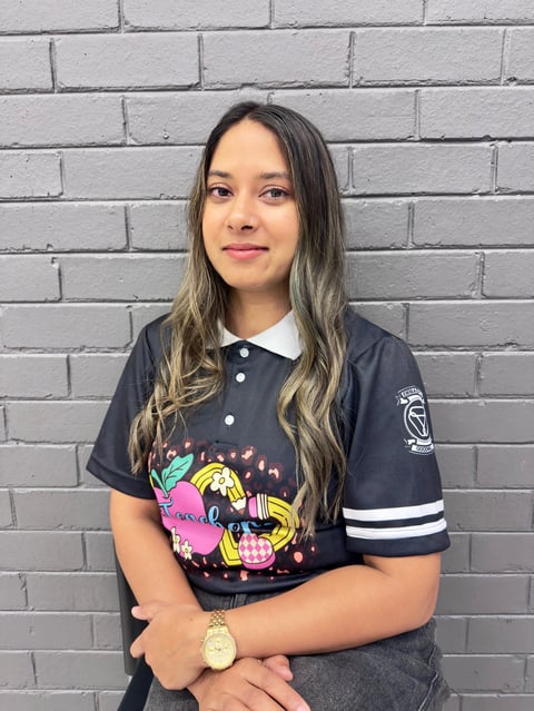 Young woman with long dark hair wearing a navy graphic t-shirt and gold watch, posing against a gray brick wall