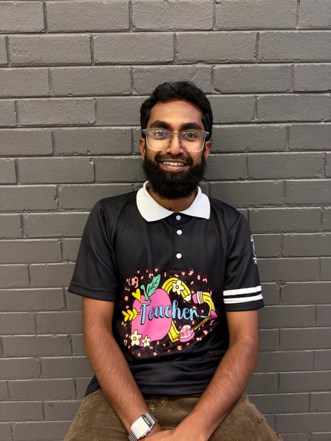 Man with glasses and beard smiling at camera wearing black polo shirt with colorful teacher graphic and white collar stripes against brick wall background