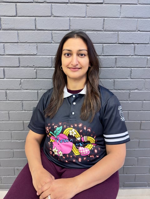 Woman with long dark hair smiling at camera, wearing dark polo shirt with colorful graphic design against gray brick wall