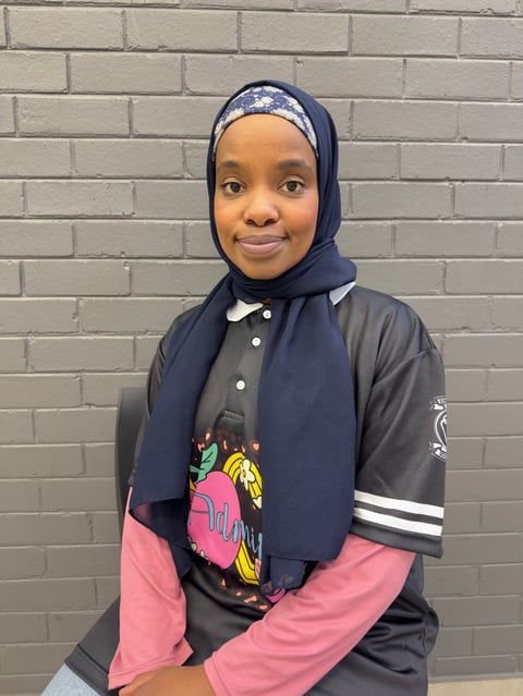 Young girl wearing a blue hijab and colorful graphic jacket smiling at the camera against a brick wall