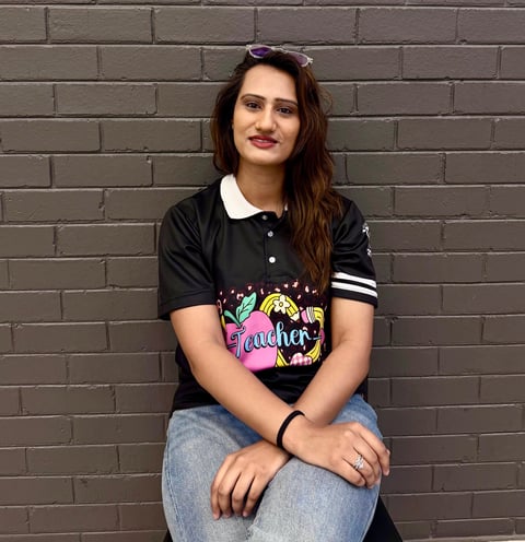 Woman with dark hair and sunglasses on head sitting against a brick wall, wearing a black polo shirt with colorful graphic design and jeans