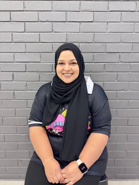 Woman wearing black hijab and dark clothing smiling at camera against white brick wall