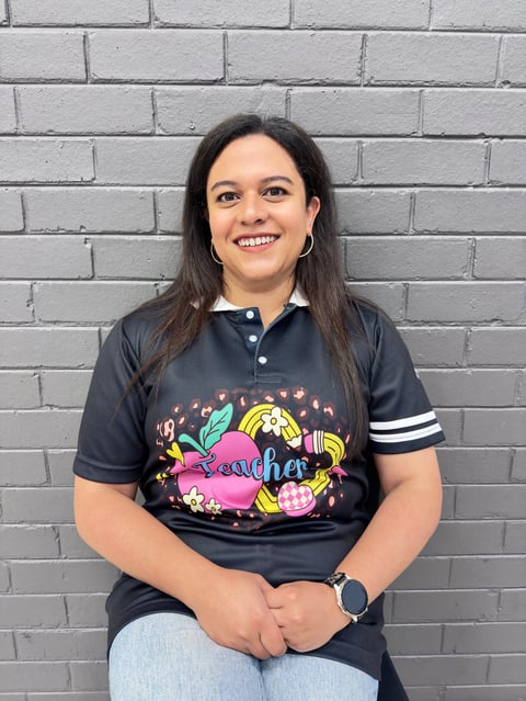 Woman smiling at camera wearing dark polo shirt with colorful teacher graphic and white stripe sleeve, standing against white brick wall