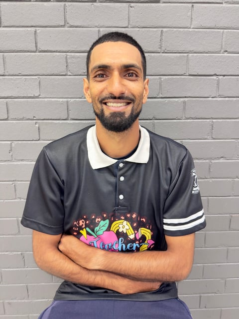 Portrait of a smiling man with a beard wearing a dark polo shirt with colorful graphics against a white brick wall