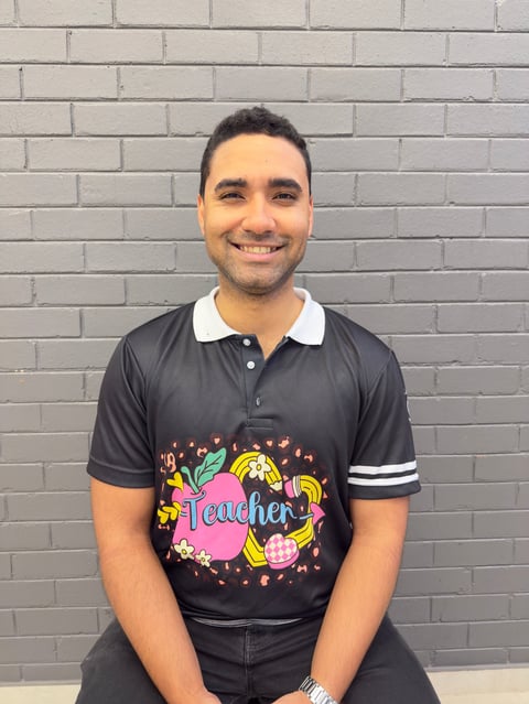 Smiling man wearing black polo shirt with colorful teacher graphic and white striped sleeves, standing in front of gray brick wall
