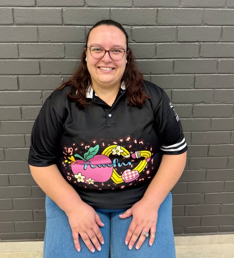 Person with long brown hair and glasses smiling at camera, wearing a black graphic t-shirt and jeans against a brick wall