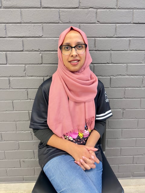 Woman wearing pink hijab and glasses sitting against a gray brick wall, smiling at camera