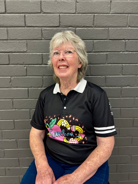 Smiling woman with white hair and glasses wearing a black polo shirt with colorful graphic design against a brick wall