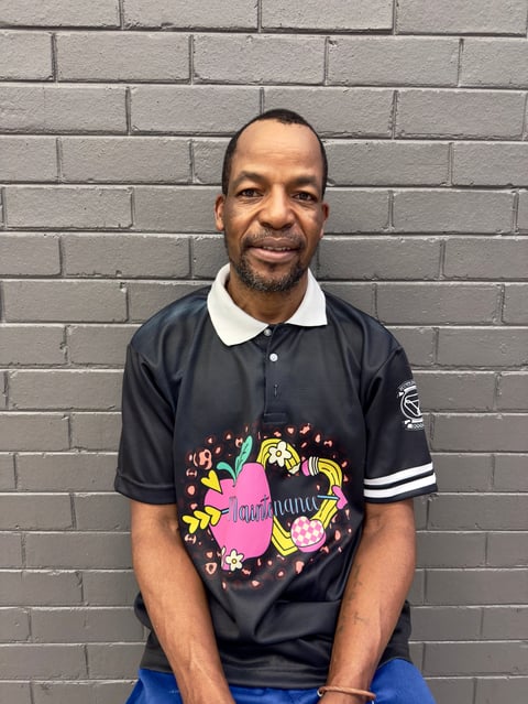 Man in black polo shirt with colorful graphic design, smiling at camera against gray brick wall background