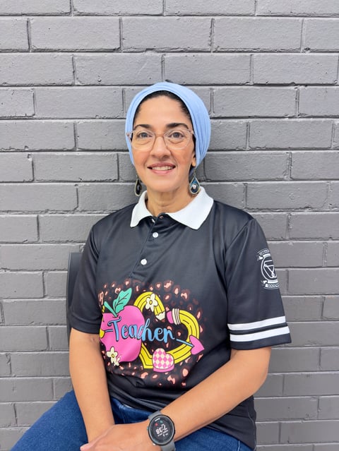 Young woman smiling at camera wearing light blue hijab, glasses, black polo shirt with colorful Teacher graphic, and watch against brick wall
